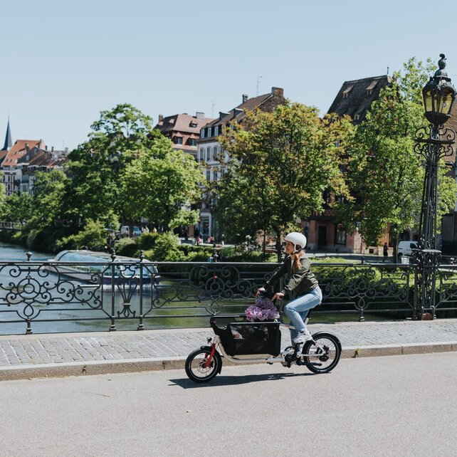 Een vrouw met fietshelm gebruikt een voorgeladen Cargo eBike voor vervoer in de stad en fietst over een brug met smeedijzeren hekken. Op de achtergrond zie je een schilderachtig decor van huizen.