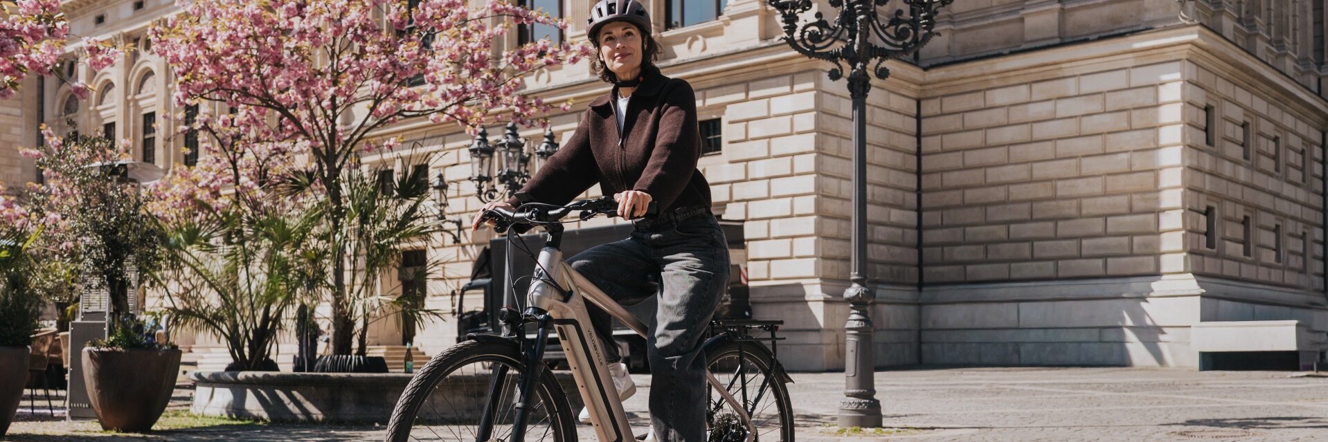Eine Frau mit Helm fährt auf einem modernen eBike über einen sonnigen Platz mit Kopfsteinpflaster. Im Hintergrund sind ein historisches Gebäude und rosa blühende Bäume zu sehen.