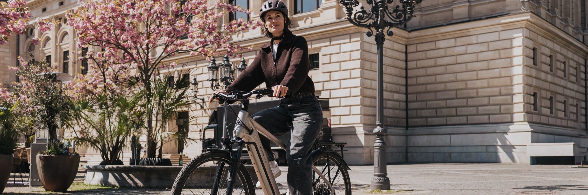 Eine Frau mit Helm fährt auf einem modernen eBike über einen sonnigen Platz mit Kopfsteinpflaster. Im Hintergrund sind ein historisches Gebäude und rosa blühende Bäume zu sehen.