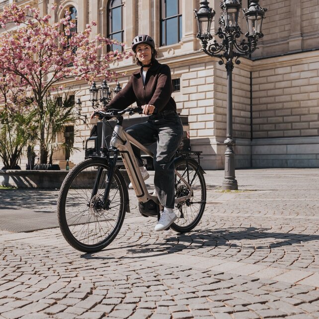 Eine Frau mit Helm fährt auf einem modernen eBike über einen sonnigen Platz mit Kopfsteinpflaster. Im Hintergrund sind ein historisches Gebäude und rosa blühende Bäume zu sehen.