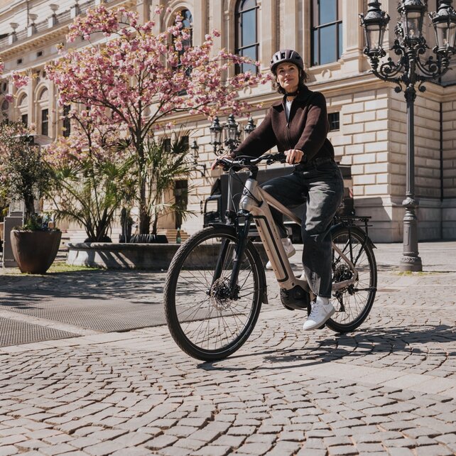 Une femme portant un casque circule sur un VAE moderne à travers une place ensoleillée ornée de pavés. À l’arrière-plan, un bâtiment historique et des arbres à fleurs roses sont visibles.