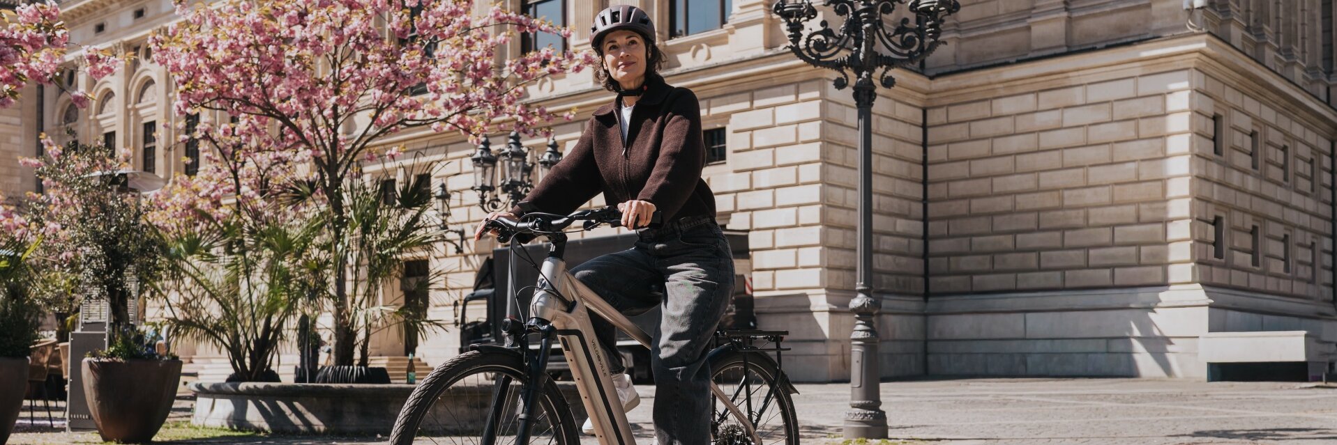 Une femme portant un casque circule sur un VAE moderne à travers une place ensoleillée ornée de pavés. À l’arrière-plan, un bâtiment historique et des arbres à fleurs roses sont visibles.