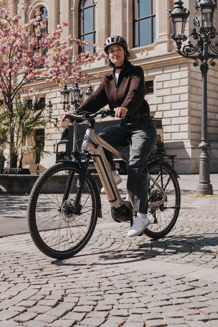 Eine Frau mit Helm fährt auf einem modernen eBike über einen sonnigen Platz mit Kopfsteinpflaster. Im Hintergrund sind ein historisches Gebäude und rosa blühende Bäume zu sehen.