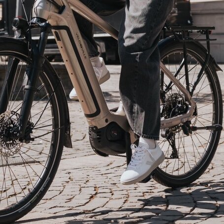 Eine Frau mit Helm fährt auf einem modernen eBike über einen sonnigen Platz mit Kopfsteinpflaster. Im Hintergrund sind ein historisches Gebäude und rosa blühende Bäume zu sehen.