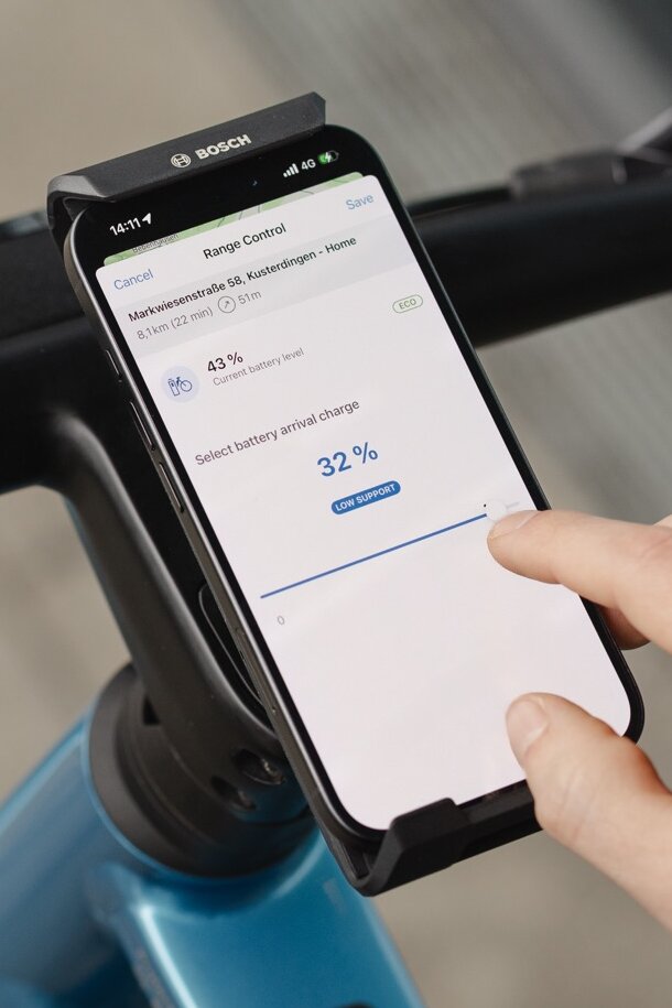 Close-up of a smartphone displaying the Range Control feature in the eBike Flow app. A user adjusts the slider to set the desired battery level to 32 percent upon arrival at their destination, optimizing the range for their planned route.