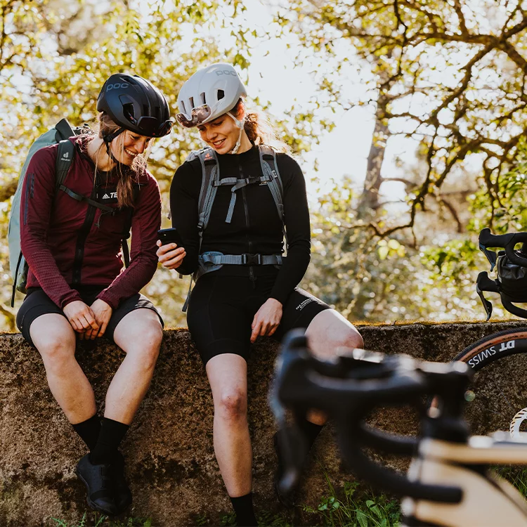 Twee vrouwen met helmen zitten op een muur en kijken samen naar een smartphone. Op de voorgrond is een e-bike te zien, terwijl op de achtergrond bomen staan.