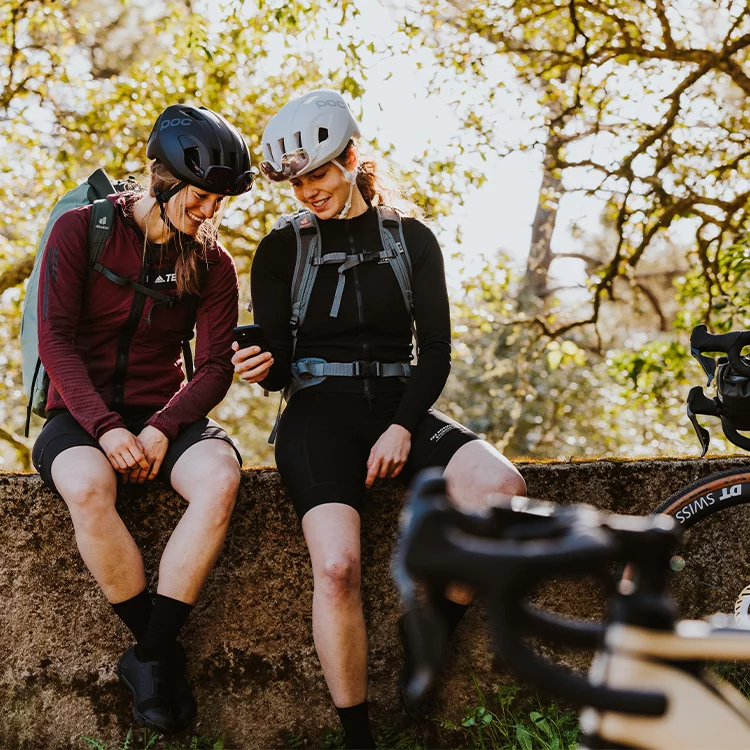 Two women with helmets sit on a wall and look at a phone together. In the foreground you can see an eBike, in the background trees.