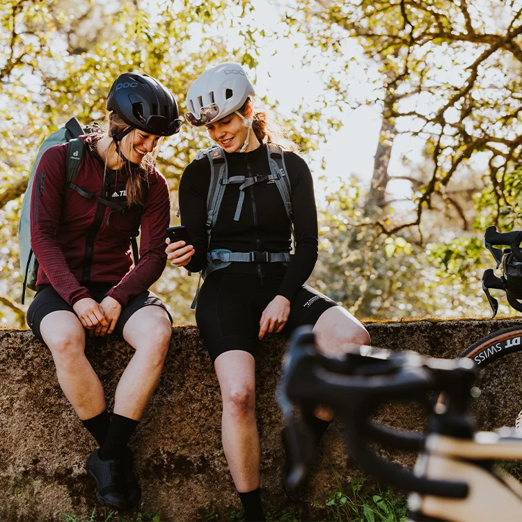 Zwei Frauen mit Helm sitzen auf einer Mauer und schauen gemeinsam auf ein Smartphone. Im Vordergrund erkennt man ein eBike, im Hintergrund Bäume.