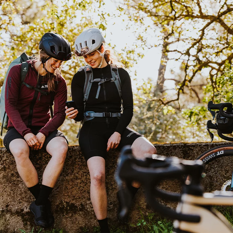 Dos mujeres con cascos se sientan en una pared y miran juntas a un teléfono. En primer plano se puede ver un eBike, en el fondo árboles.