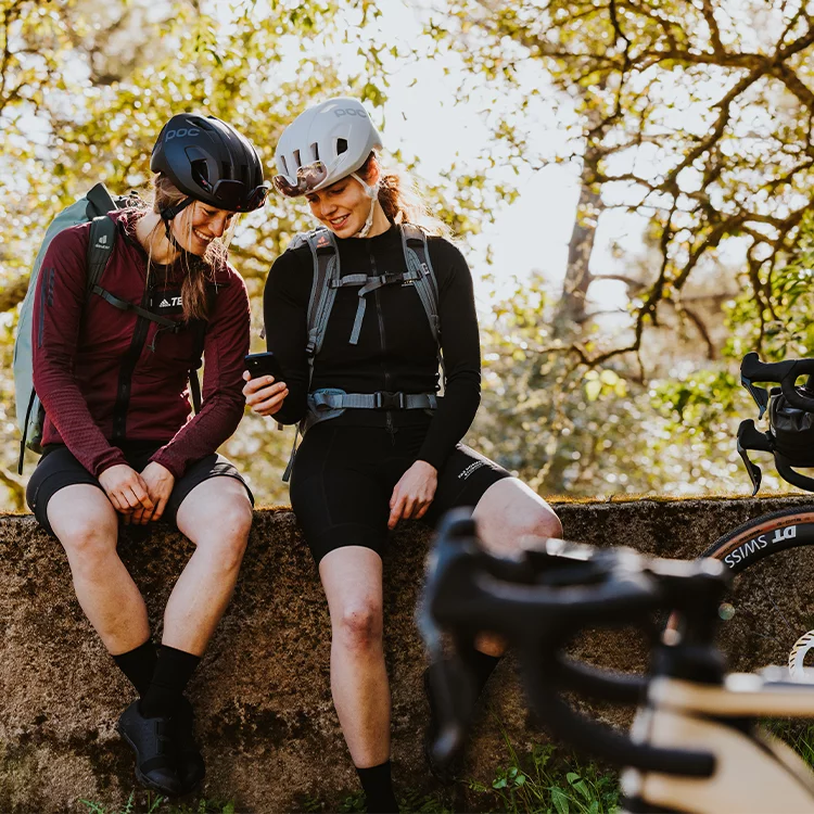 Two women with helmets sit on a wall and look at a phone together. In the foreground you can see an eBike, in the background trees.