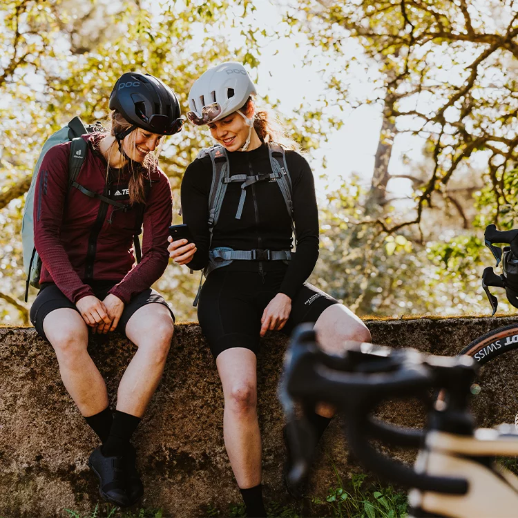 Two women with helmets sit on a wall and look at a phone together. In the foreground you can see an eBike, in the background trees.