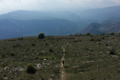 Un Vététiste parcourt un long sentier de terre à travers une étendue verte et dégagée. À l’arrière-plan, un paysage montagneux recouvert de végétation disparaît dans la brume.