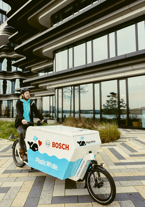 Een man rijdt op een Cargo Bike langs een weg naast een gebouw met grote glazen gevels. De laadruimte van de bakfiets is uitgebouwd en bedrukt in blauw en wit, met een zwarte walvis, het rode Bosch-logo en "PlasticWhale" erop.