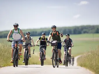 A group of cyclists is riding up a country lane, with one rider showing the "peace" sign. In the background, there is a beautiful green landscape.