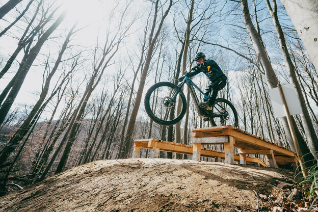 Ein Mann springt auf einem blauen eMountainbike von einer Holzrampe hinunter. Im Vordergrund sieht man den erdigen Hügel, auf den der Fahrradfahrer springt, im Hintergrund kahle hohe Bäume und blauen Himmel.