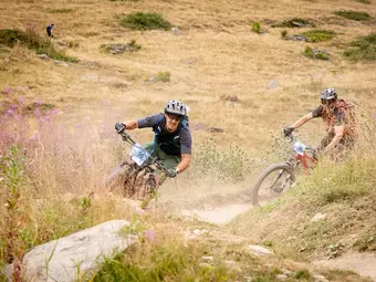 Two mountain bikers are riding up a narrow trail and leaning into the curve.