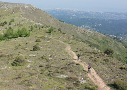 Deux Vététiste roulent l’un derrière l’autre sur un sentier en terre qui longe la crête d’une montagne. Vue sur un paysage vallonné à l’horizon.