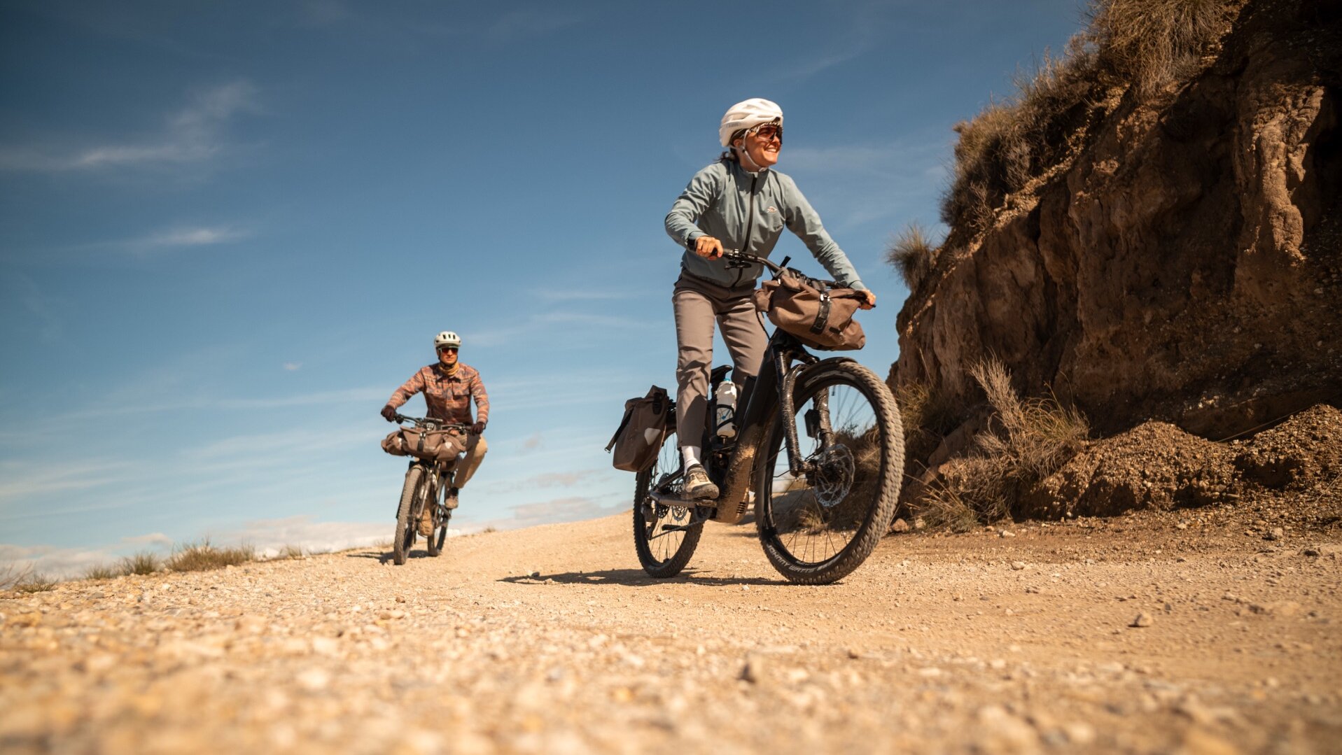 Two people, a woman in the foreground and a man further back, are riding their eBikes along a gravel path. Both wear helmets, and their bikes are packed with bags for a bike tour. The scene unfolds on a sunny day in a hilly, dry landscape.