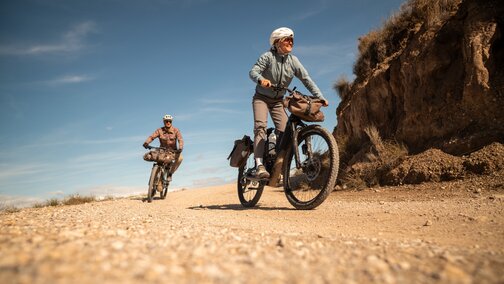 Two people, a woman in the foreground and a man further back, are riding their eBikes along a gravel path. Both wear helmets, and their bikes are packed with bags for a bike tour. The scene unfolds on a sunny day in a hilly, dry landscape.