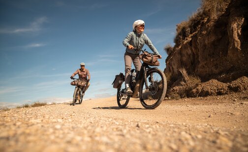 Two people, a woman in the foreground and a man further back, are riding their eBikes along a gravel path. Both wear helmets, and their bikes are packed with bags for a bike tour. The scene unfolds on a sunny day in a hilly, dry landscape.