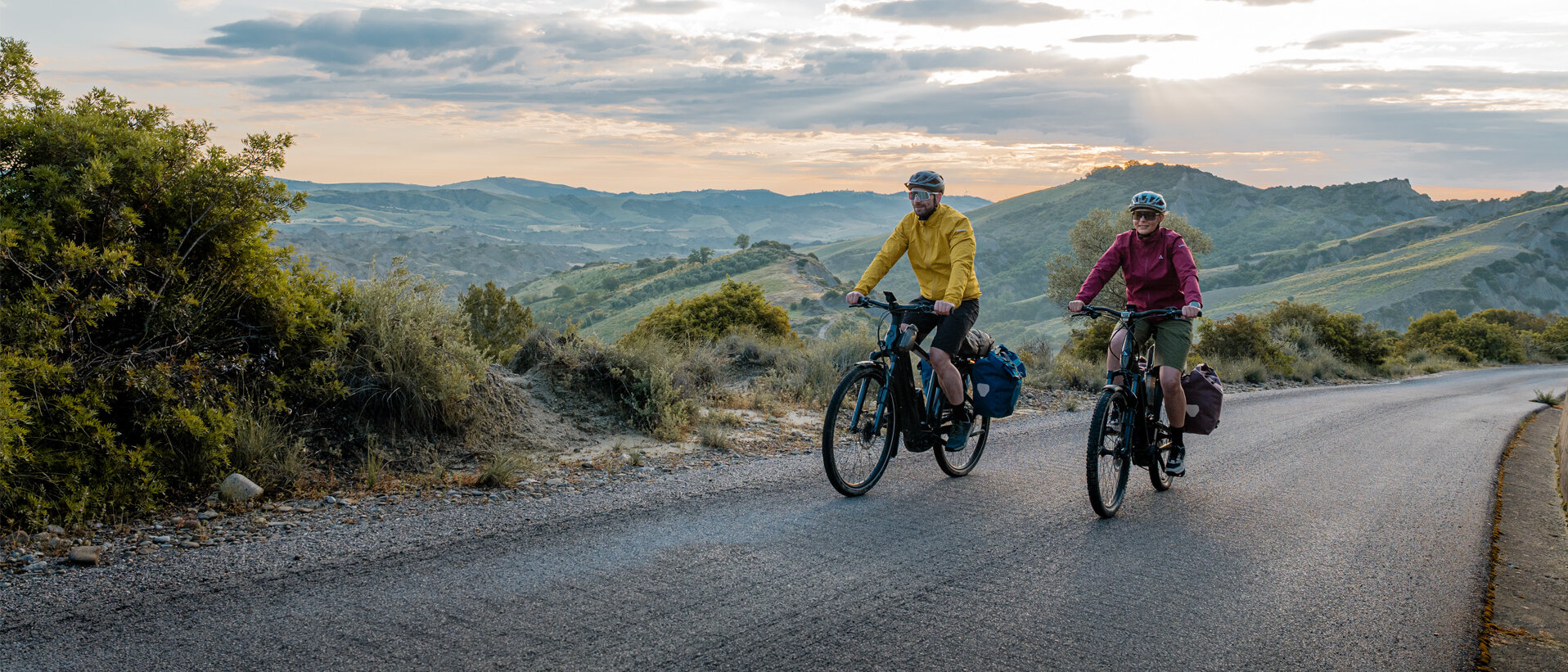 Eine Frau und ein Mann fahren mit ihren eTrekking-Bikes auf einer Straße, im Hintergrund ist eine weitläufige Landschaft zu sehen.