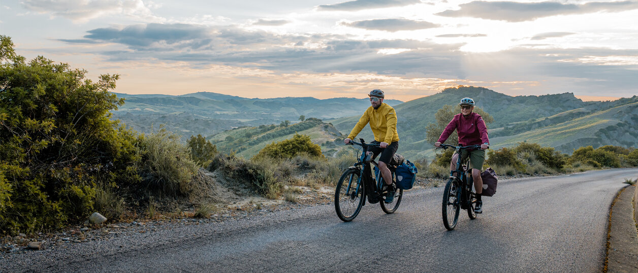 Eine Frau und ein Mann fahren mit ihren eTrekking-Bikes auf einer Straße, im Hintergrund ist eine weitläufige Landschaft zu sehen.