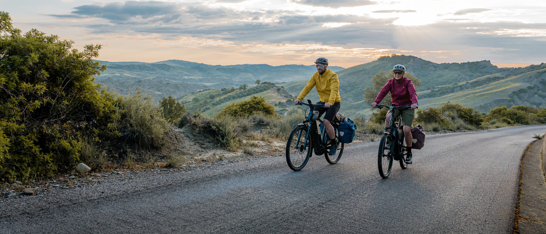 Eine Frau und ein Mann fahren mit ihren eTrekking-Bikes auf einer Straße, im Hintergrund ist eine weitläufige Landschaft zu sehen.
