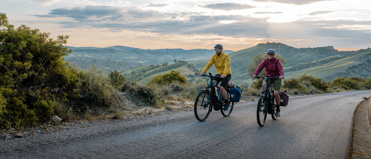 Eine Frau und ein Mann fahren mit ihren eTrekking-Bikes auf einer Straße, im Hintergrund ist eine weitläufige Landschaft zu sehen.