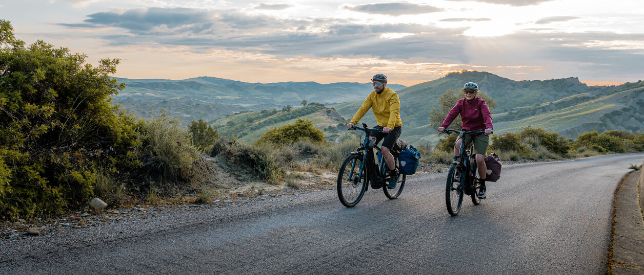 Eine Frau und ein Mann fahren mit ihren eTrekking-Bikes auf einer Straße, im Hintergrund ist eine weitläufige Landschaft zu sehen.