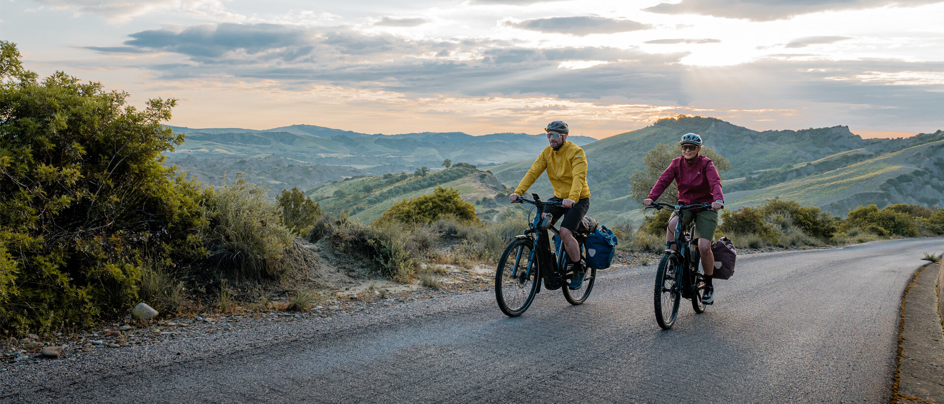 Eine Frau und ein Mann fahren mit ihren eTrekking-Bikes auf einer Straße, im Hintergrund ist eine weitläufige Landschaft zu sehen.