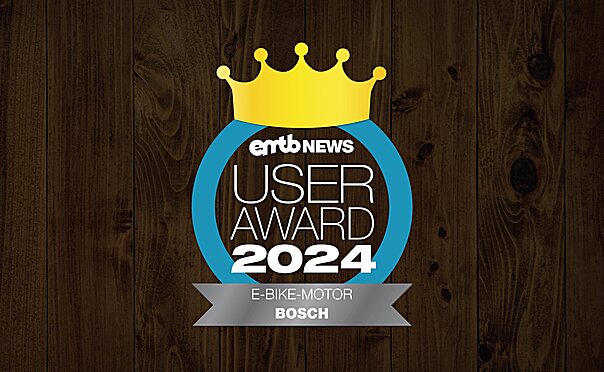 Icon with the text "eMTB NEWS USER AWARD 2024": A golden crown above the text, enclosed by a blue circle. "E-BIKE-MOTOR BOSCH" is written at the bottom. The background has a dark wood finish.