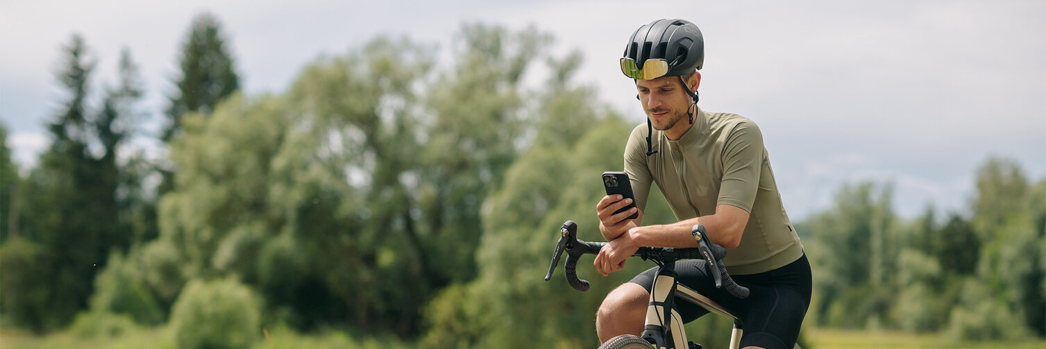 Een man maakt een pauze op zijn e-bike en kijkt op zijn smartphone. Op de achtergrond zijn weiden en bossen te zien.