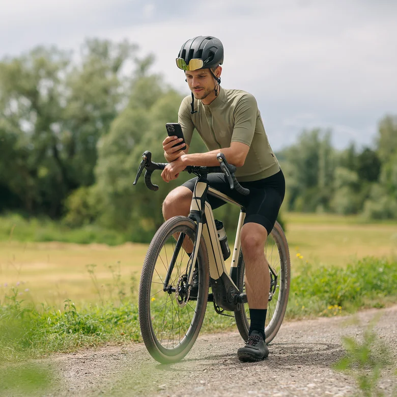 A man takes a break on his eBike, looking at his smartphone. In the background, meadows and forests are visible.