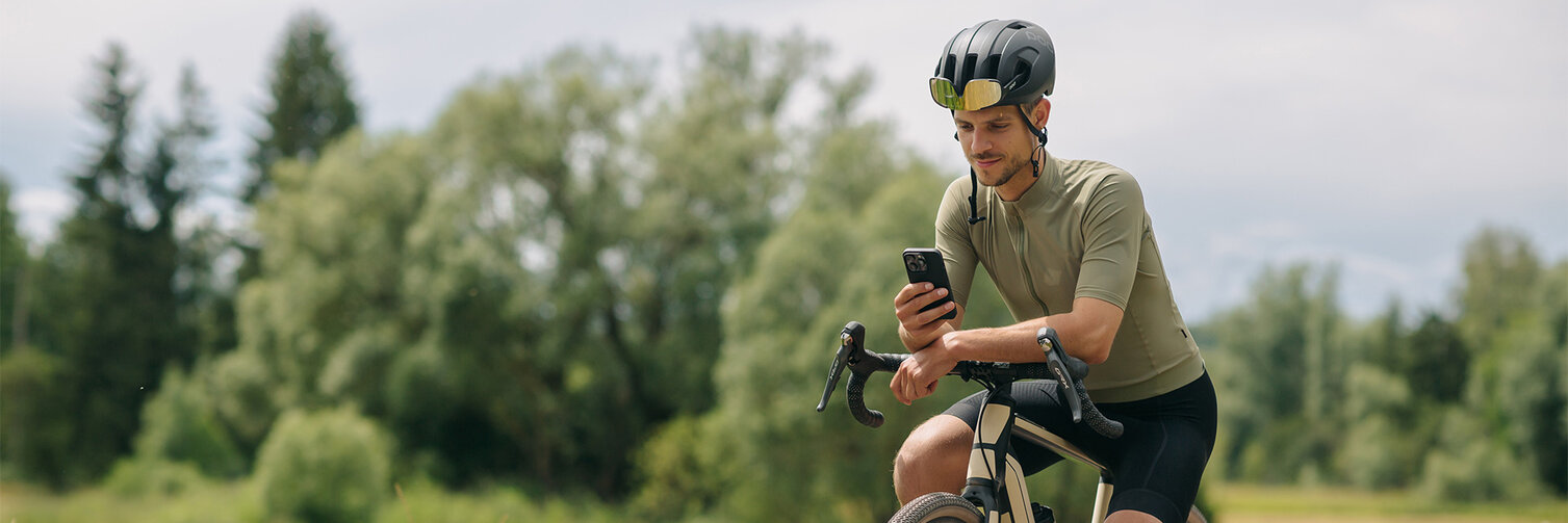 En man tar en paus på sin elcykel och tittar på sin smartphone. I bakgrunden syns ängar och skog.