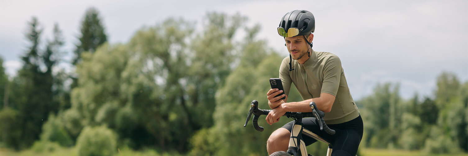 A man takes a break on his eBike, looking at his smartphone. In the background, meadows and forests are visible.