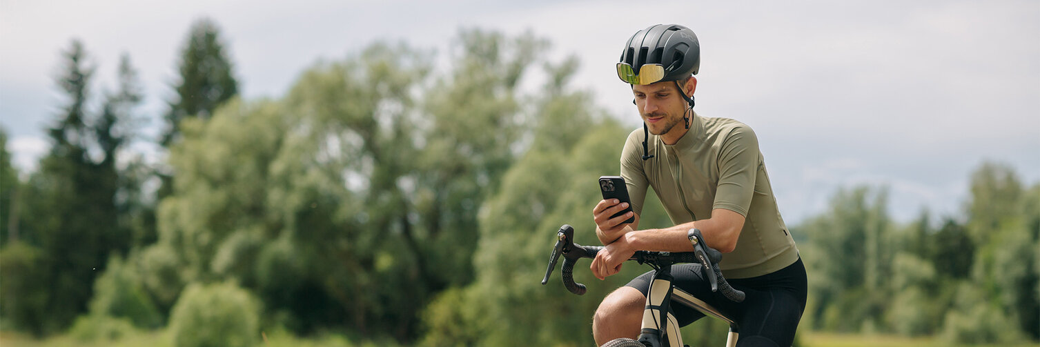 A man takes a break on his eBike, looking at his smartphone. In the background, meadows and forests are visible.