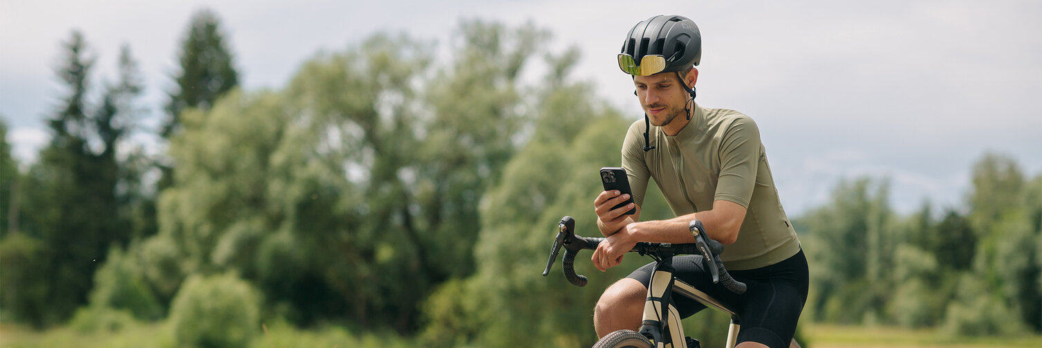Un homme fait une pause sur son vélo électrique, consulte son smartphone. À l'arrière-plan, on peut voir des prairies et des forêts.