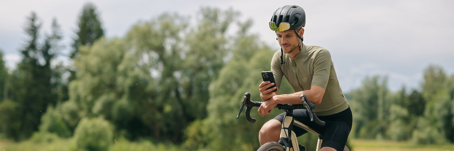 Un homme fait une pause sur son vélo électrique, consulte son smartphone. À l'arrière-plan, on peut voir des prairies et des forêts.