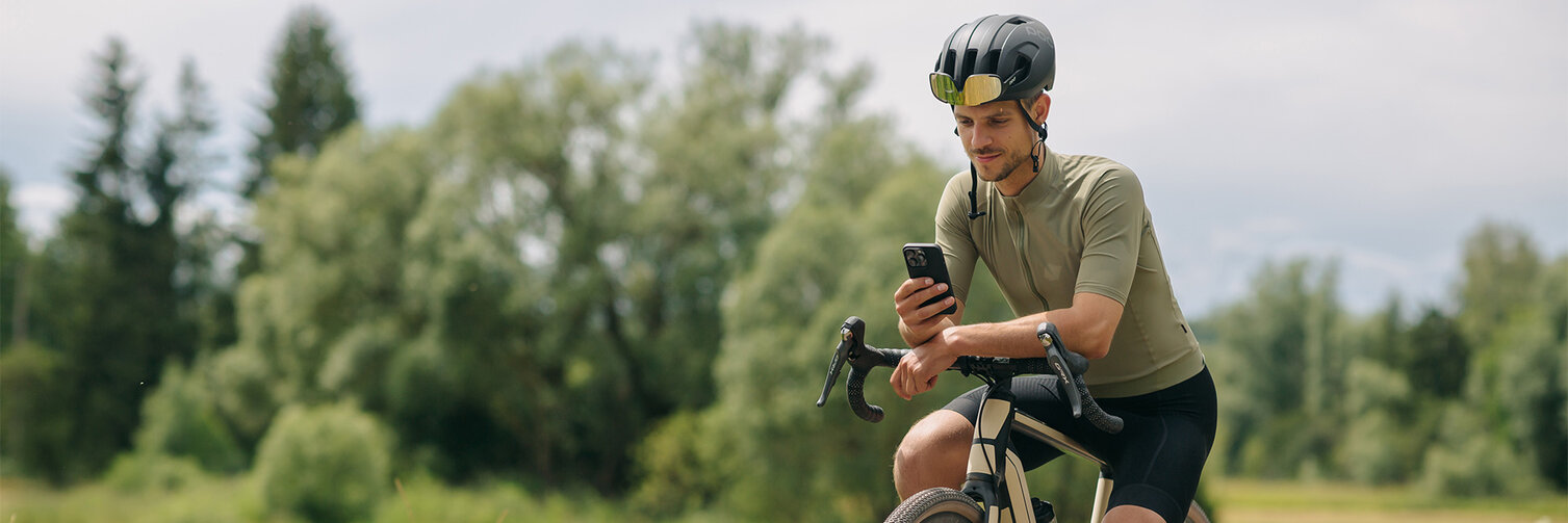 A man takes a break on his eBike, looking at his smartphone. In the background, meadows and forests are visible.