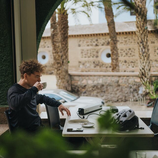Ein Mountainbiker macht eine Pause auf der Terrasse eines Cafés und trinkt aus einer Tasse. Auf dem Tisch vor ihm lädt der Akku seines eBikes, daneben liegt sein Fahrradhelm. Durch die Fensterbögen sind Palmen und eine mediterrane Steinmauer zu sehen.