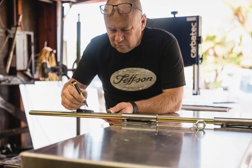 A man wearing a top labeled 'Jeffson' marks a pole with a pen in a workshop.