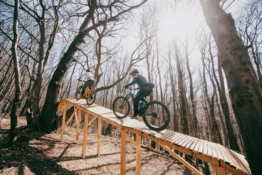 Zwei Personen fahren hintereinander auf eMountainbikes eine dünne, durch einen Wald führende Holzbrücke entlang. Die Bäume des Waldes sind kahl, am Boden liegt Laub.