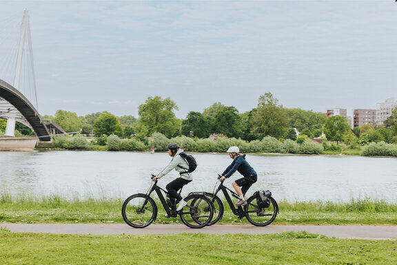 Zwei Personen genießen eine eBike-Tour auf einem Radweg entlang eines Flusses. Die städtische Kulisse mit einer modernen Brücke im Hintergrund unterstreicht die Kombination aus Freizeit und urbaner Mobilität.