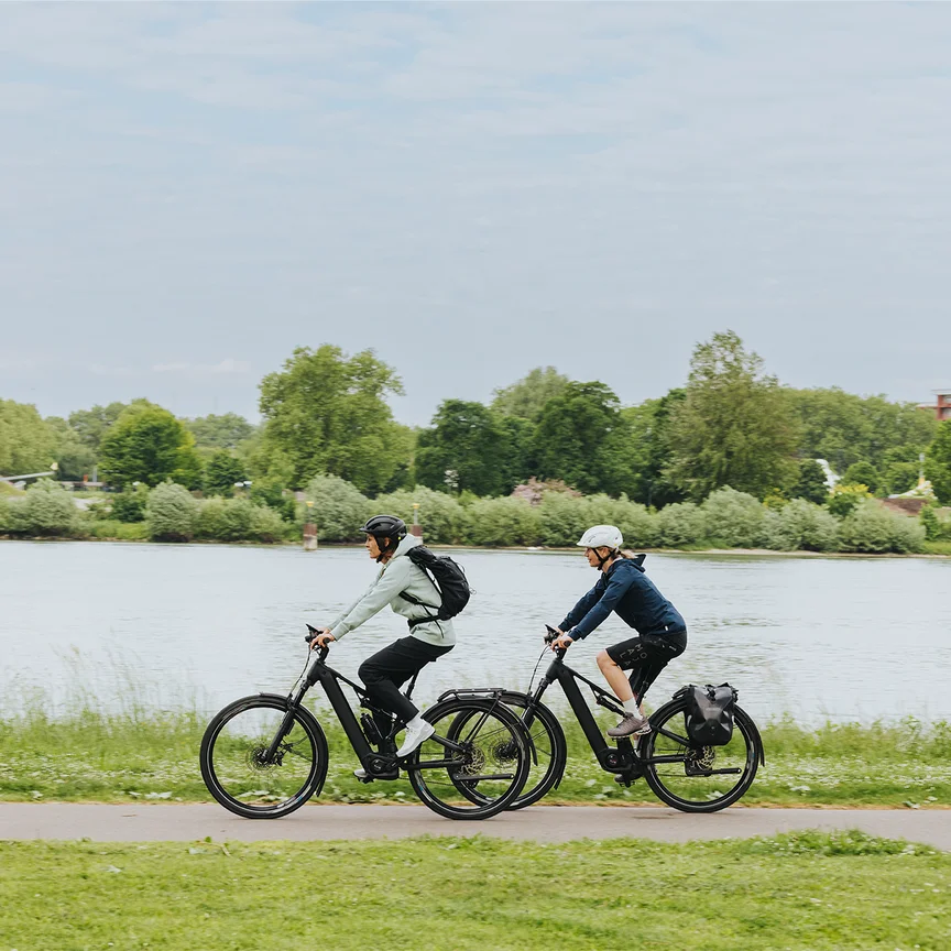 Två personer njuter av en elcykeltur på en cykelväg längs en flod. Den urbana bakgrunden med en modern bro i bakgrunden framhäver kombinationen av fritid och urban mobilitet.