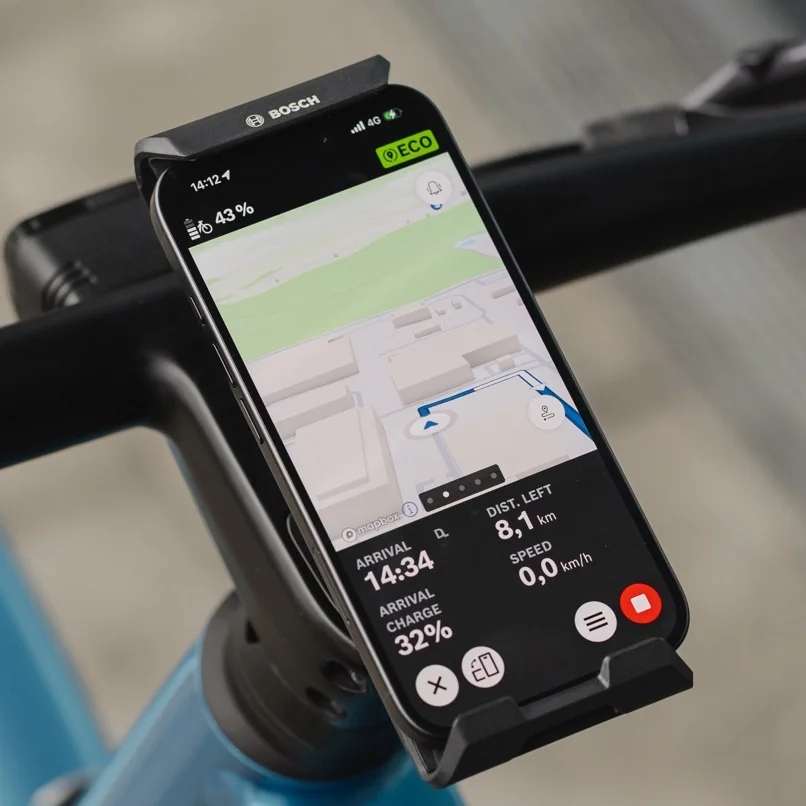 Närbild av en smartphone som är monterad på styret i Bosch-elcykel SmartphoneGrip Vertical och används som elcykel display för navigering. eBike Flow-app visar en färgglad karta med en blå ruttlinje, en kommande sväng åt vänster samt återstående avstånd och ankomsttid längst ner på skärmen.