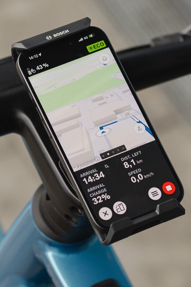 Zbliżenie smartfona przymocowanego do kierownicy w Bosch rower ze wspomaganiem elektrycznym / e-rower SmartphoneGrip Vertical i używanego jako rower ze wspomaganiem elektrycznym / e-rower wyświetlacz do nawigacja. Aplikacja eBike Flow pokazuje kolorową mapę drogową z niebieską linią trasy, nadchodzący skręt w lewo oraz pozostałą odległość i czas przybycia na dole ekranu.