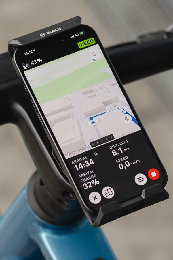 Närbild av en smartphone som är monterad på styret i Bosch-elcykel SmartphoneGrip Vertical och används som elcykel display för navigering. eBike Flow-app visar en färgglad karta med en blå ruttlinje, en kommande sväng åt vänster samt återstående avstånd och ankomsttid längst ner på skärmen.