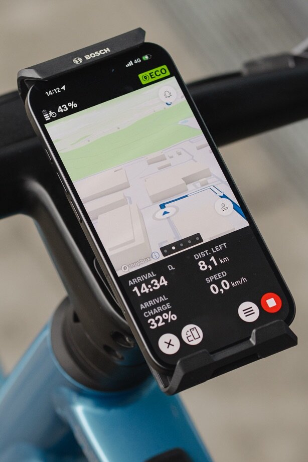 Primo piano di uno smartphone fissato al manubrio nel eBike SmartphoneGrip Vertical Bosch e utilizzato come eBike display per la navigazione. L'app eBike Flow mostra una mappa stradale colorata con una linea blu del percorso, una svolta imminente a sinistra e la distanza rimanente e l'orario di arrivo nella parte inferiore dello schermo.