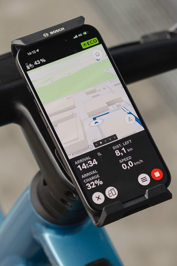 Primer plano de un smartphone fijado al manillar en el eBike SmartphoneGrip Vertical Bosch, utilizado como eBike display para navegación. La aplicación eBike Flow muestra un mapa de carreteras a color con una línea de ruta azul, una indicación de giro próximo a la izquierda, así como la distancia restante y el tiempo de llegada en la parte inferior de la pantalla.
