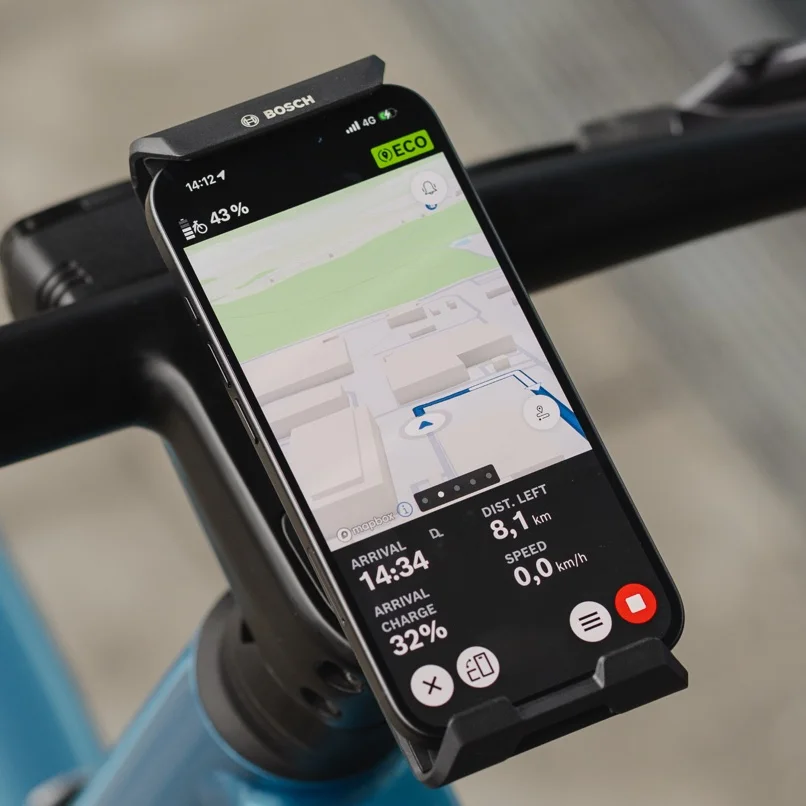 Close-up of a smartphone attached to the handlebars in the Bosch eBike SmartphoneGrip Vertical, used as an eBike display for navigation. The eBike Flow app shows a colorful road map with a blue route line, an upcoming left turn, and the remaining distance and arrival time at the bottom of the screen.